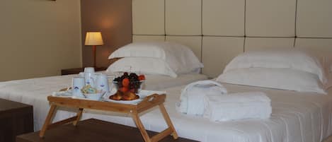 A hotel room with two beds, a wooden side table with a tray of fruit and beverages, and a lamp on the nightstand.