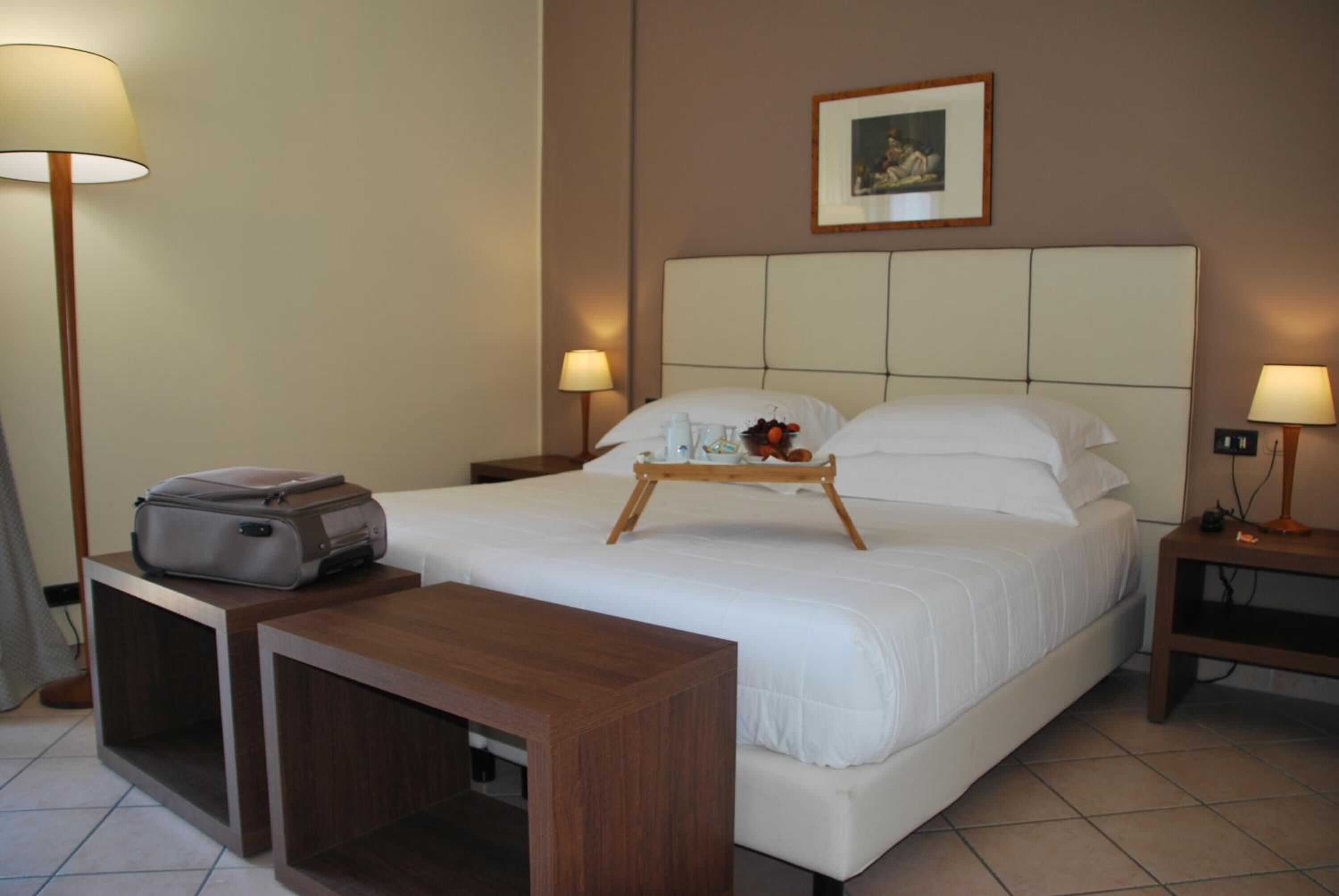 Una habitación de hotel con una cama, lámparas de noche, una mesita de noche con lámpara, una pequeña mesa con una bandeja de fruta y una maleta en el suelo.
