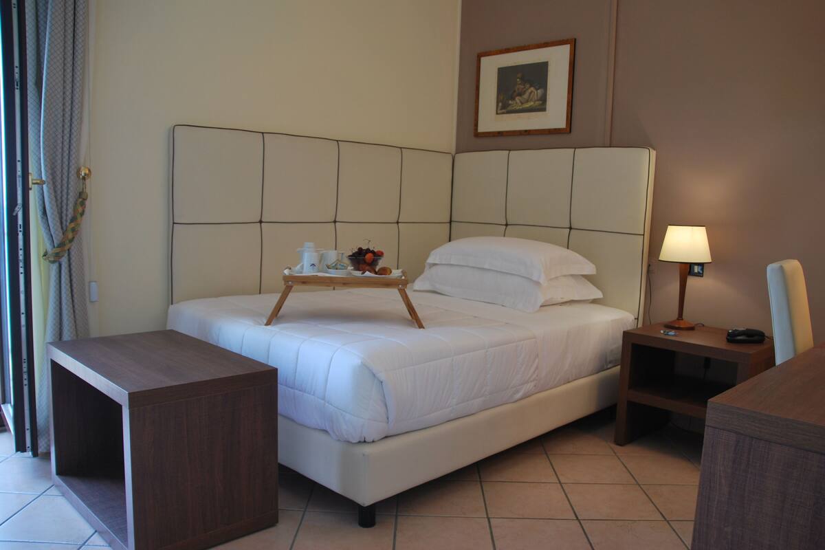 A hotel room with a bed, bedside table, lamp, and a small wooden side table with a tray of items.