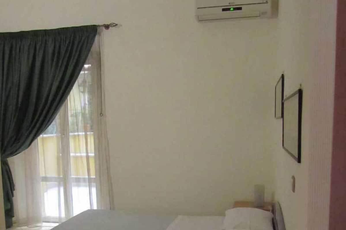 A bedroom with a bed, a nightstand, an air conditioning unit, and a window with curtains.