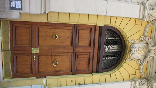 Una puerta de madera grande con un picaporte de metal, ubicada en un edificio amarillo que tiene un arco decorativo encima.