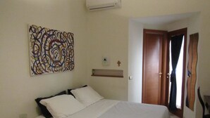 Un dormitorio con una cama, una mesita de noche, aire acondicionado instalado en la pared, una puerta de madera y una obra de arte en la pared.