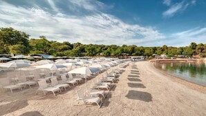 A beach with rows of white sun loungers and umbrellas, a sandy shore, and a calm body of water. - Valamar Camping Lanterna (Tar-Vabriga)