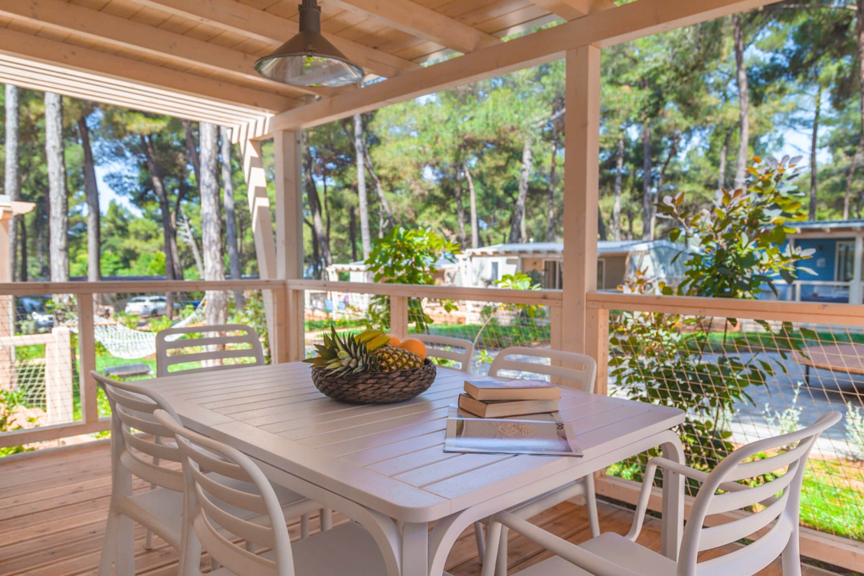 Un área de comedor al aire libre techada, con una mesa dispuesta para dos personas, sillas blancas alrededor y una cesta de fruta sobre la mesa.