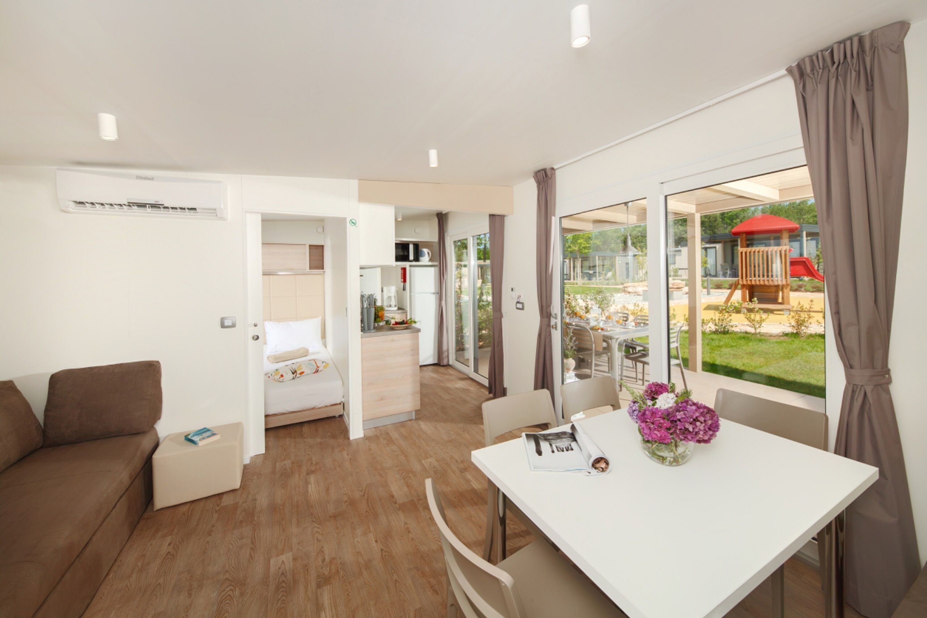 Ein modernes Hotelzimmer mit Holzboden, einem weißen Bett, einem weißen Esstisch mit Blumenvase und Blick auf einen Spielplatz.