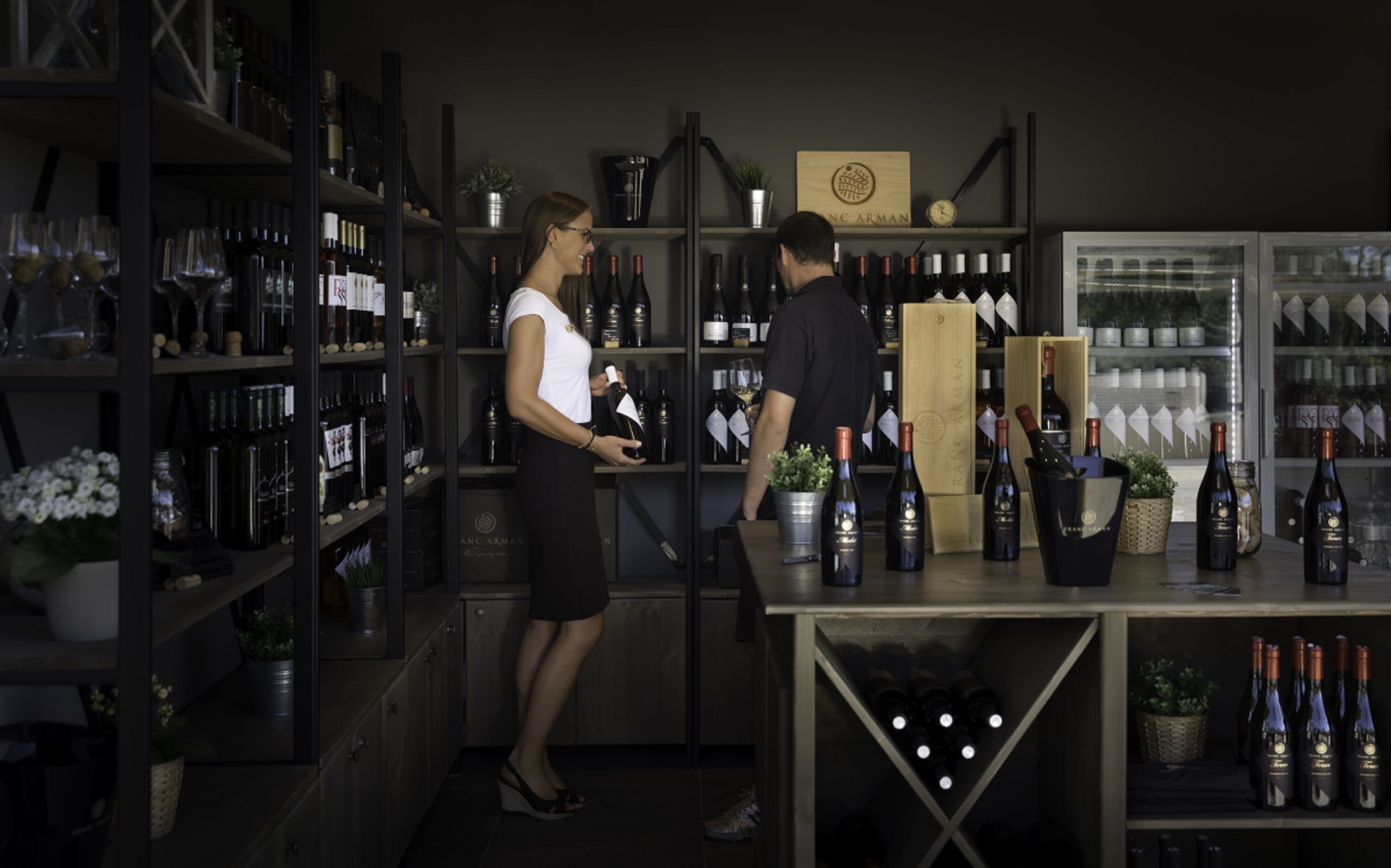 A wine cellar with shelves stocked with wine bottles and a few customers browsing.