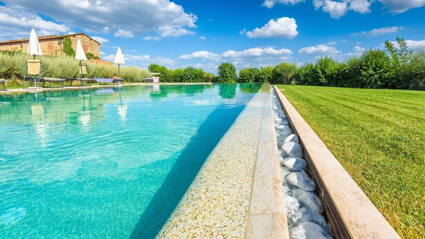 Una piscina cristalina con sillas de descanso y sombrillas, rodeada de un césped cuidado y un edificio al fondo.
