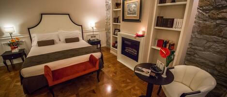 A hotel room with a large bed, a small table with a chair, a bookshelf, and a fireplace.