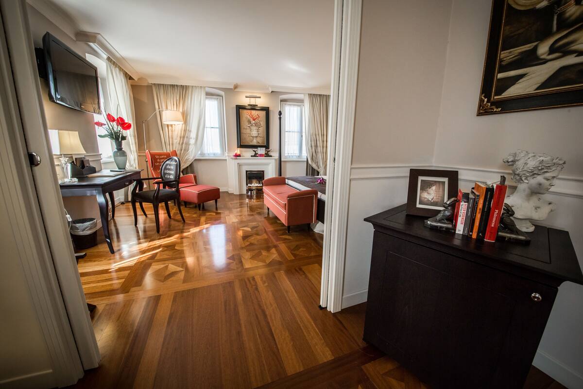A spacious room with a wooden parquet floor, a fireplace, a dining area with a red chair, and a desk with a television.