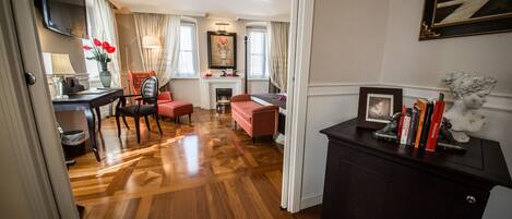 A spacious room with a wooden parquet floor, a fireplace, a dining area with a red chair, and a desk with a television.