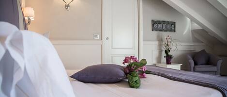 A neatly made bed with a purple pillow and a vase of flowers, a chair, and a wall-mounted light fixture in a hotel room.