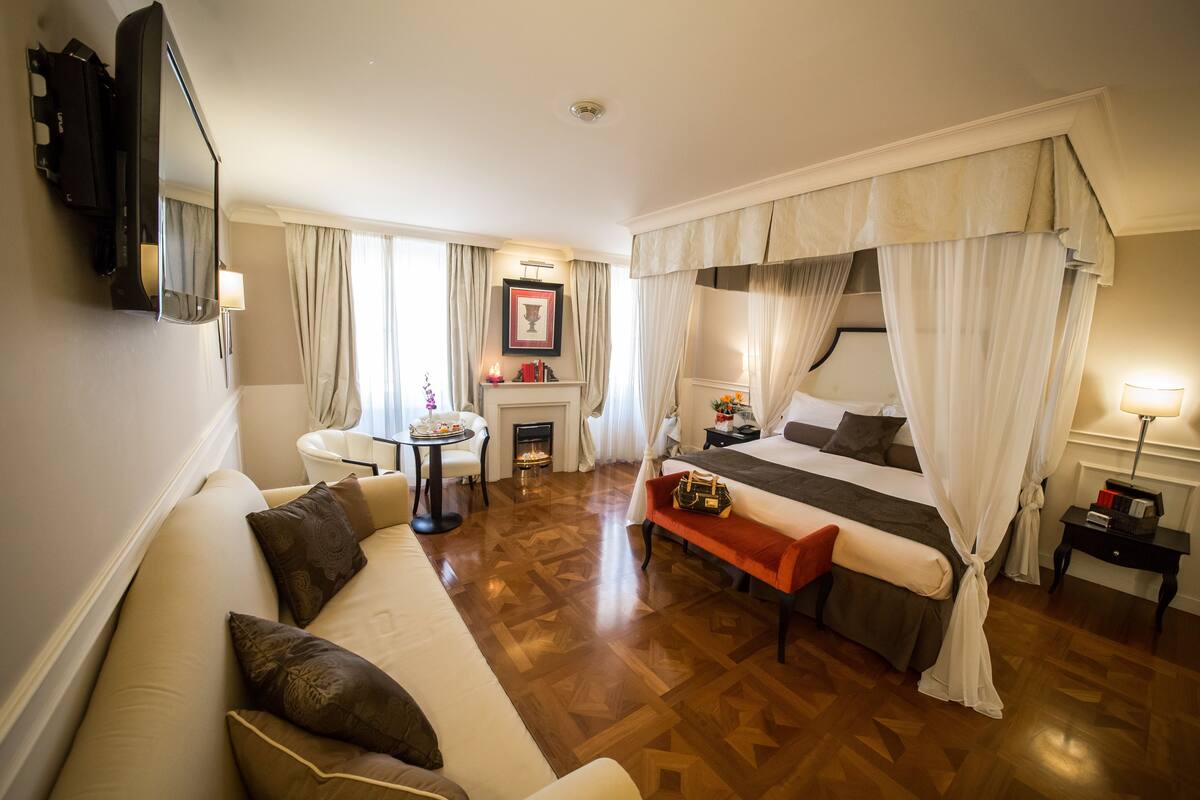 A hotel room with a bed, a sofa, a small table, a fireplace, and a television mounted on the wall.