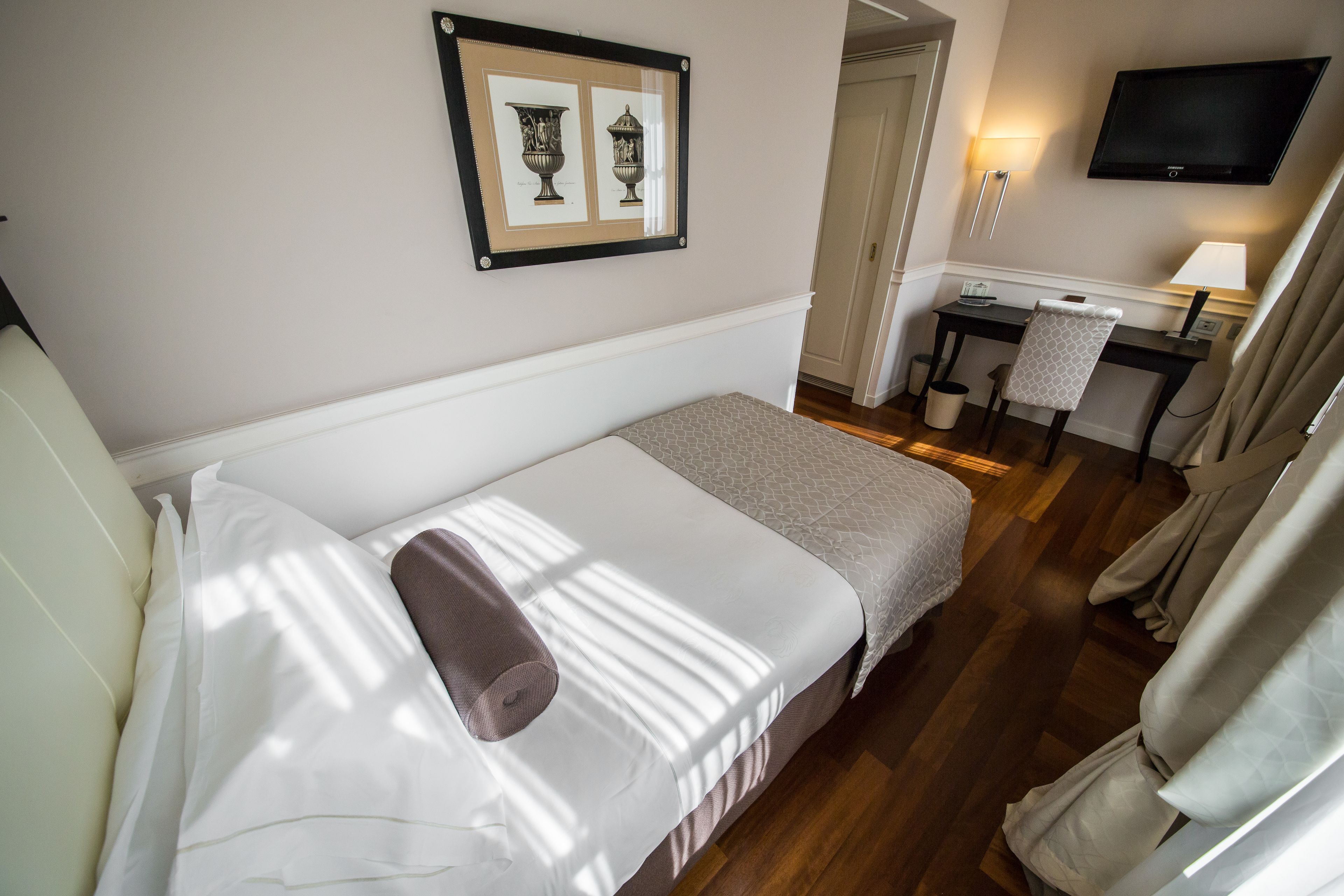 A hotel room with a bed, a desk, a chair, a TV, and framed artwork on the wall.