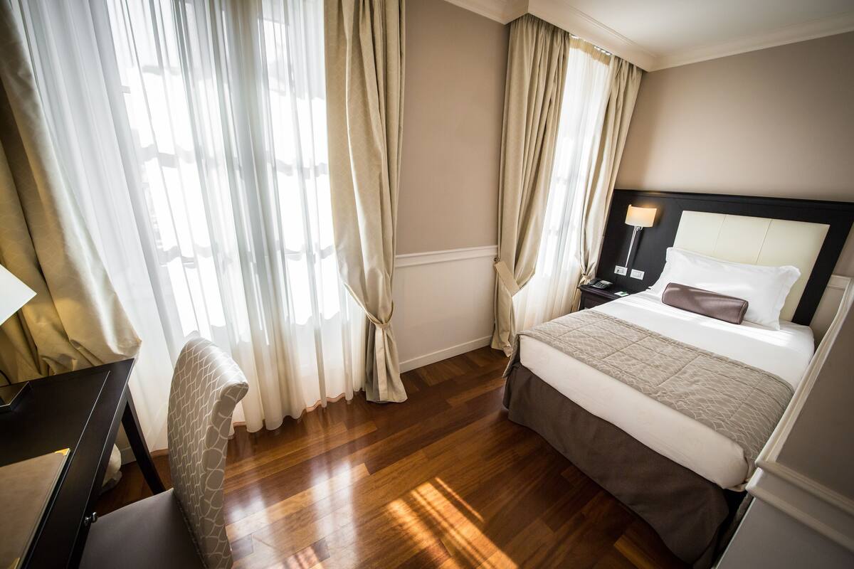 A hotel room with a bed, a desk, a chair, and a nightstand with a lamp.