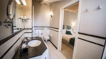 Ein Badezimmer mit Waschbecken, Spiegel und Toilette. Eine Tür führt zu einem Schlafzimmer mit Bett und Nachttisch.