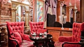 A room with a stone wall, a large mirror, red upholstered chairs, a round table with cups and saucers, and a candelabra. - Hotel Maritza (Plovdiv)