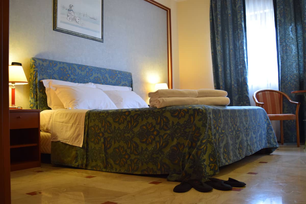 A hotel room with a bed, two bedside lamps, a chair, and a pair of slippers on the floor.
