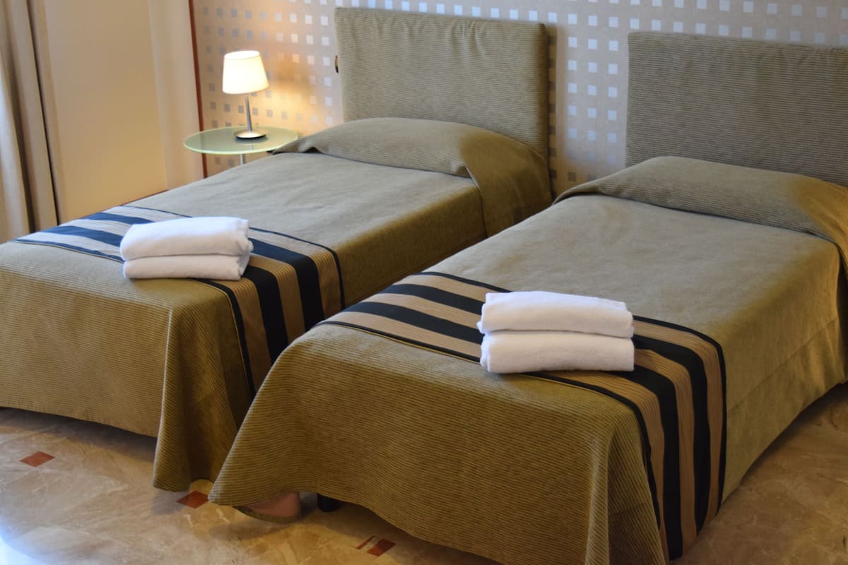 Two single beds with striped bedding and folded towels, a small table with a lamp, and a patterned wall in a hotel room.