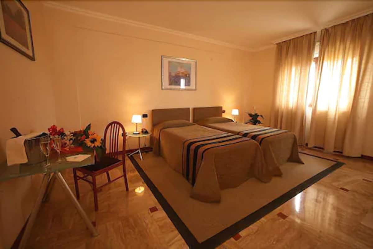 A hotel room with two beds, a desk with a chair, a small table with a vase of flowers, and a framed picture on the wall.