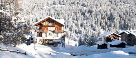 Una cabaña de madera de varios pisos con balcones, rodeada de árboles y montañas cubiertas de nieve.