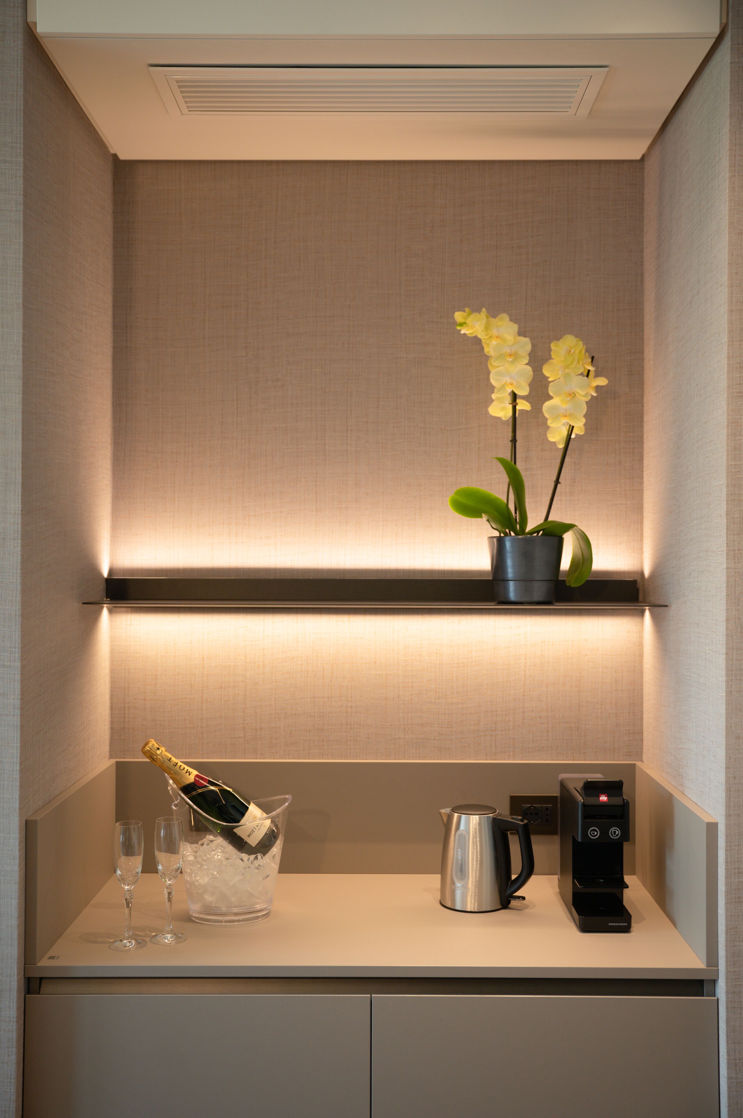 Un minibar con una botella de champán, dos copas, una cafetera y una orquídea en maceta.