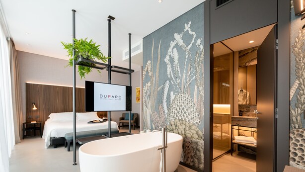 Una habitación de hotel moderna con una bañera grande, una cama, un escritorio y un televisor mostrando 'DUPARC'.