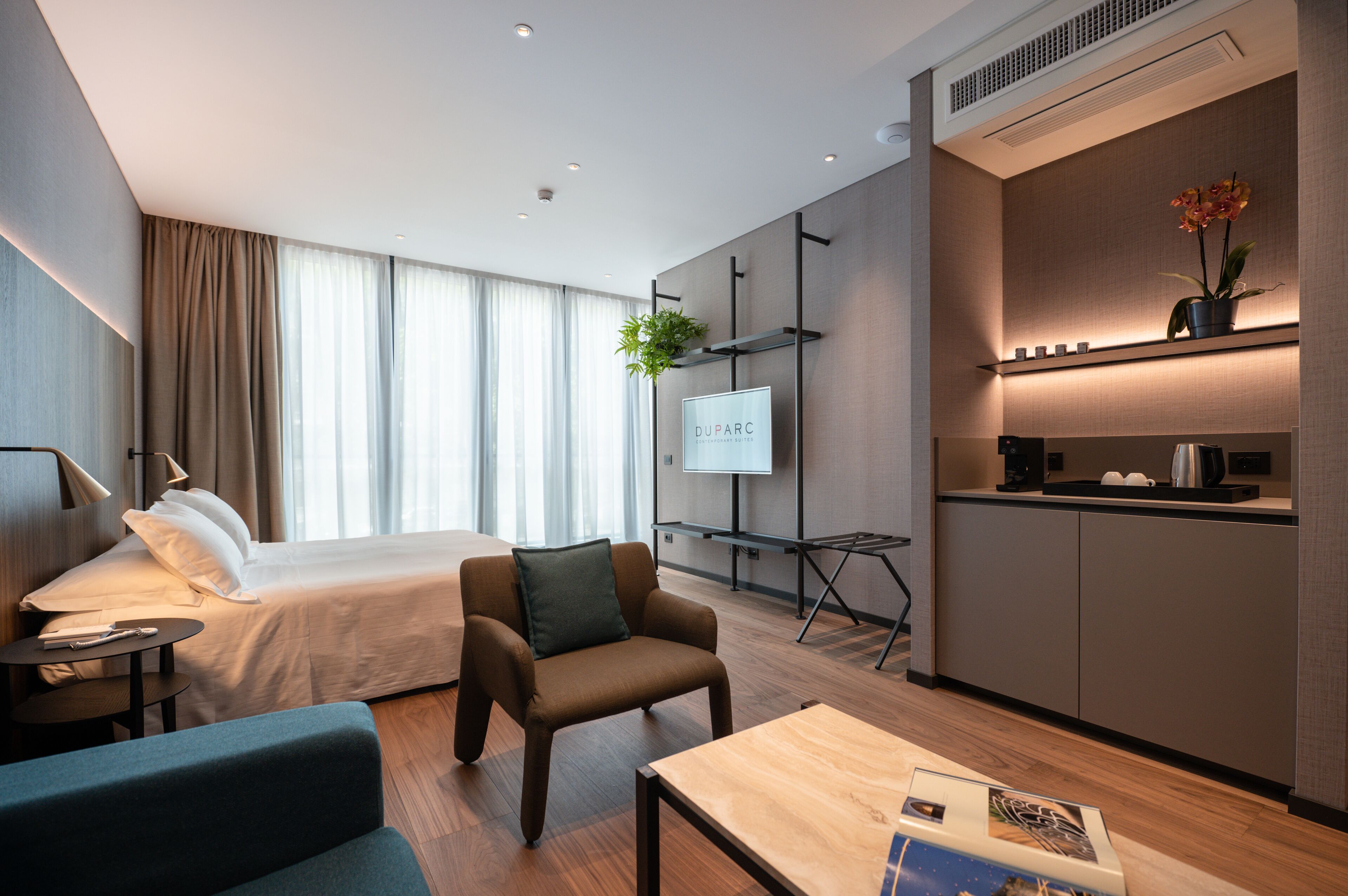 A modern hotel room with a bed, a seating area, a small table, a TV, a plant, and a kitchenette.