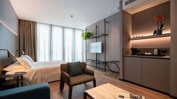 A modern hotel room with a bed, a seating area, a small table, a TV, a plant, and a kitchenette.