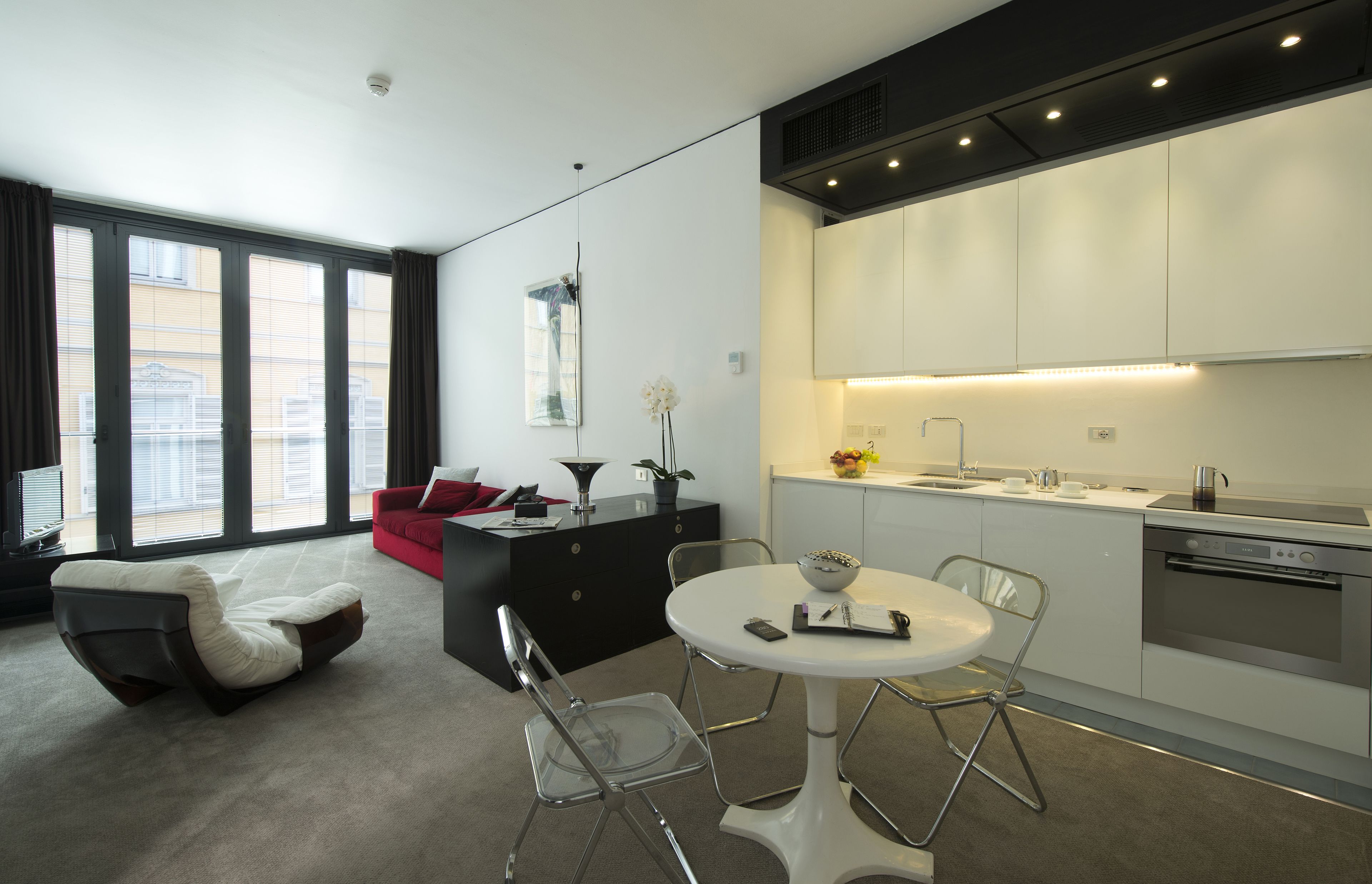 A modern living room with a sofa, a dining table with chairs, a kitchen with white cabinets, and a large window with curtains.