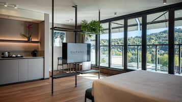 A modern hotel room with a large bed, a TV, a desk with a chair, a small kitchen area, and a balcony with a view of the city.