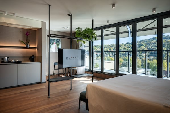A modern hotel room with a large bed, a TV, a desk with a chair, a small kitchen area, and a balcony with a view of the city.