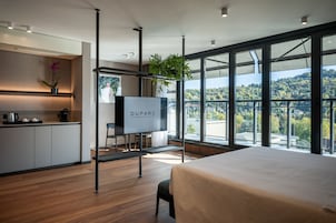 A modern hotel room with a large bed, a TV, a desk with a chair, a small kitchen area, and a balcony with a view of the city.