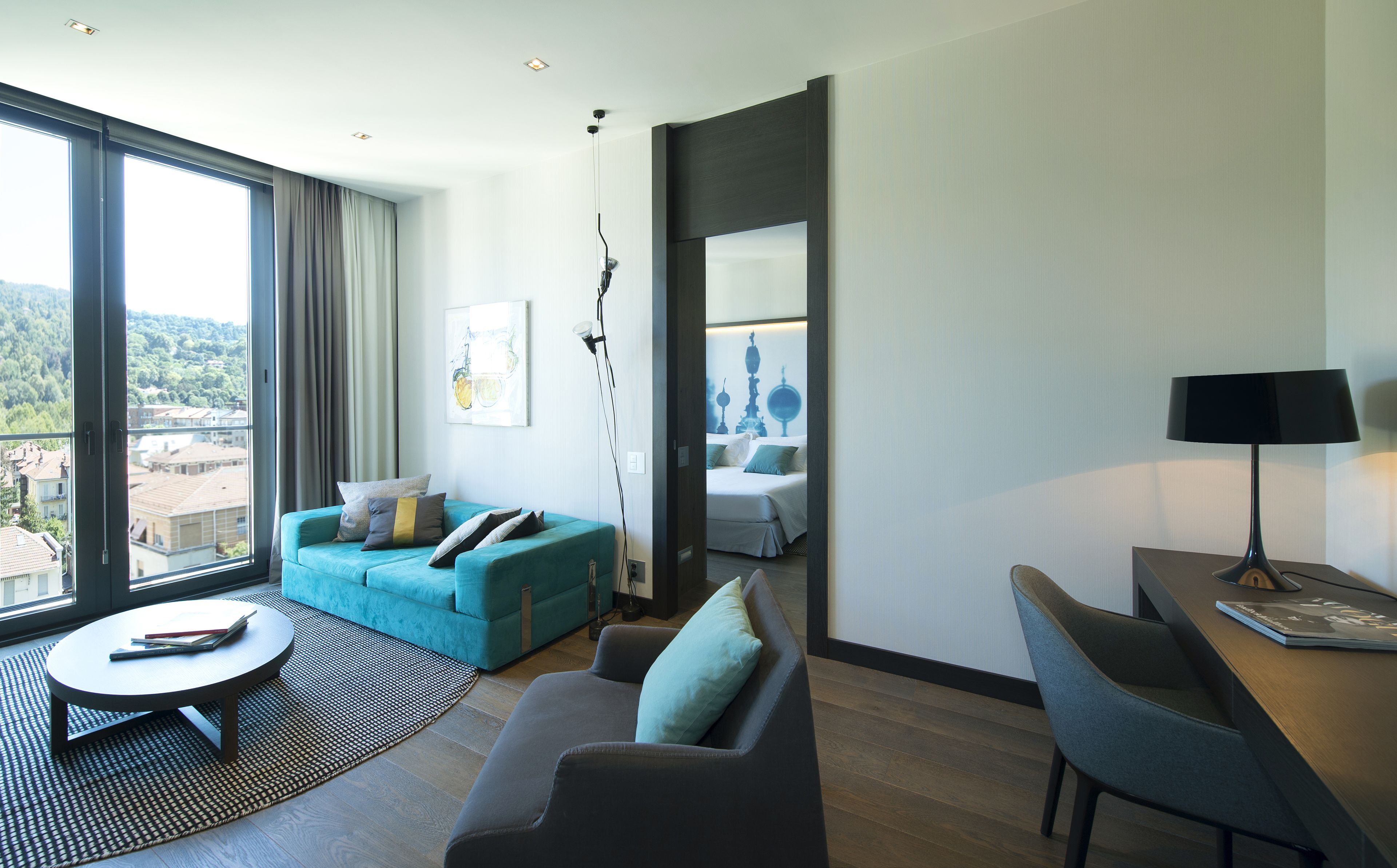 Un salon moderne avec un canapé turquoise, un fauteuil gris et une petite table ronde. Une grande fenêtre offre une vue sur les immeubles et les arbres. On aperçoit un lit avec une tête de lit blanche à travers une porte ouverte.
