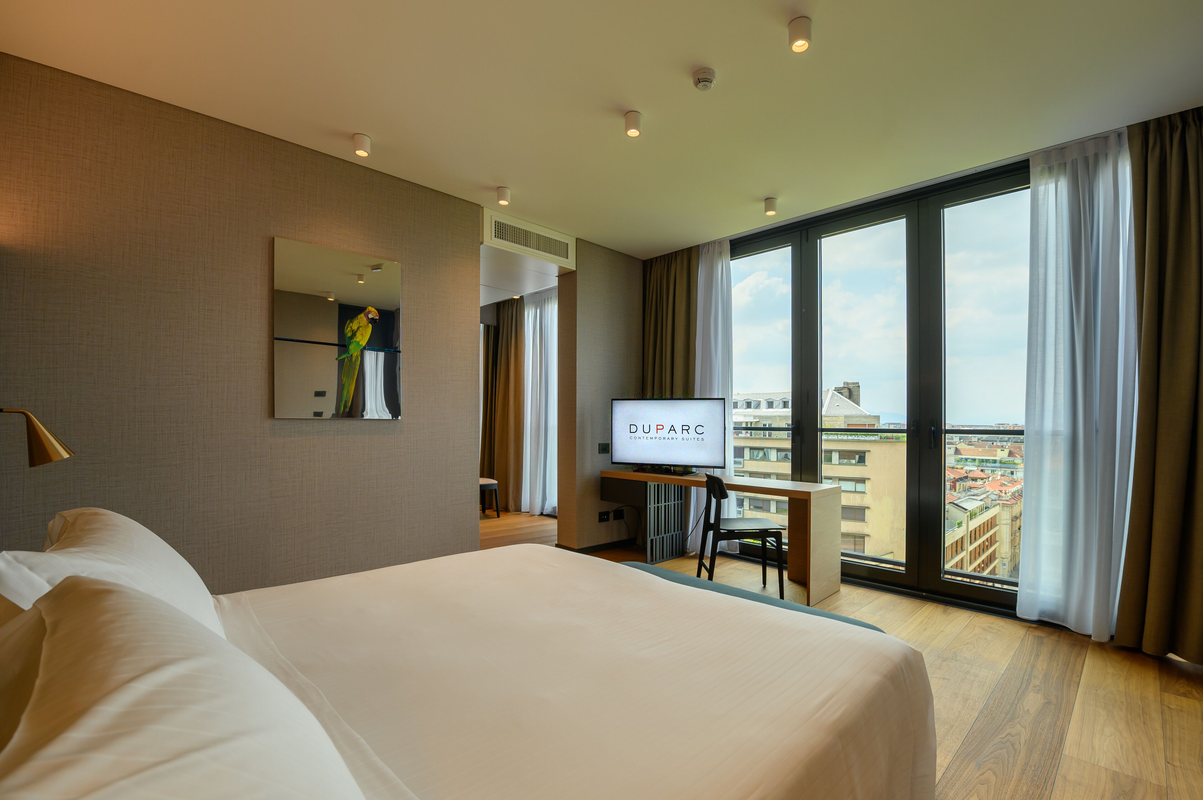 Een hotelkamer met een groot bed, een bureau met een televisie, een spiegel en uitzicht op de stad door grote ramen.