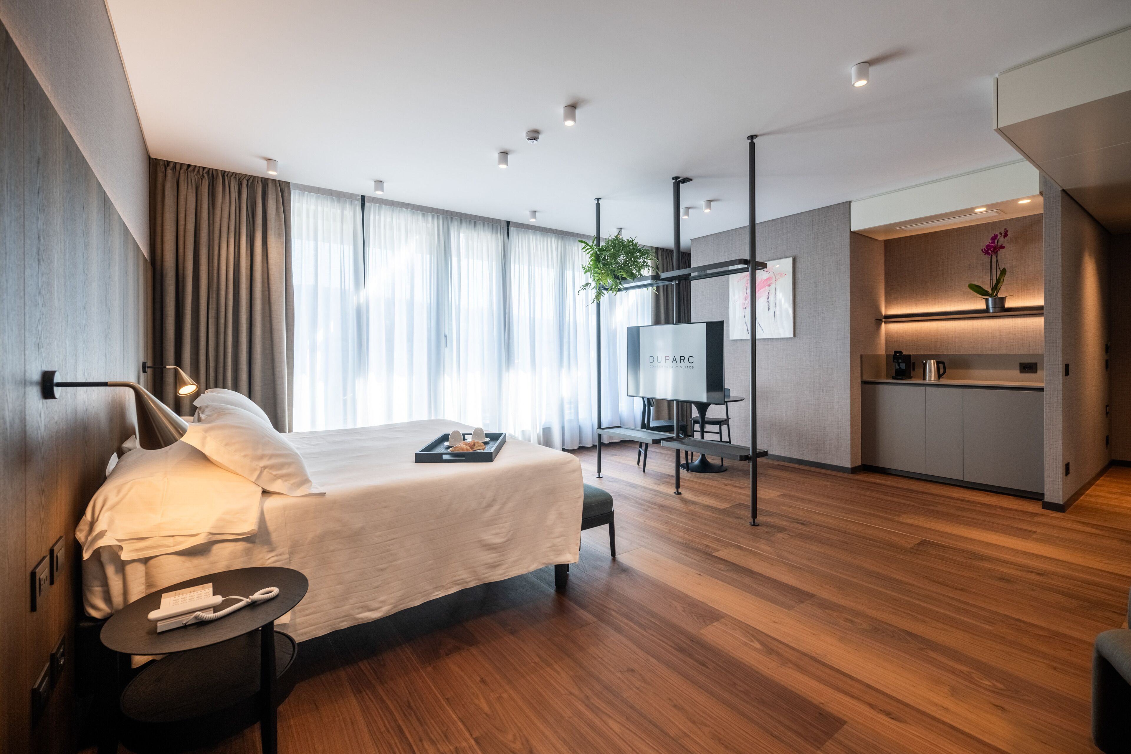 A modern hotel room with a large bed, bedside table, and a desk with a chair. There is a kitchenette area with a coffee machine and a potted plant.