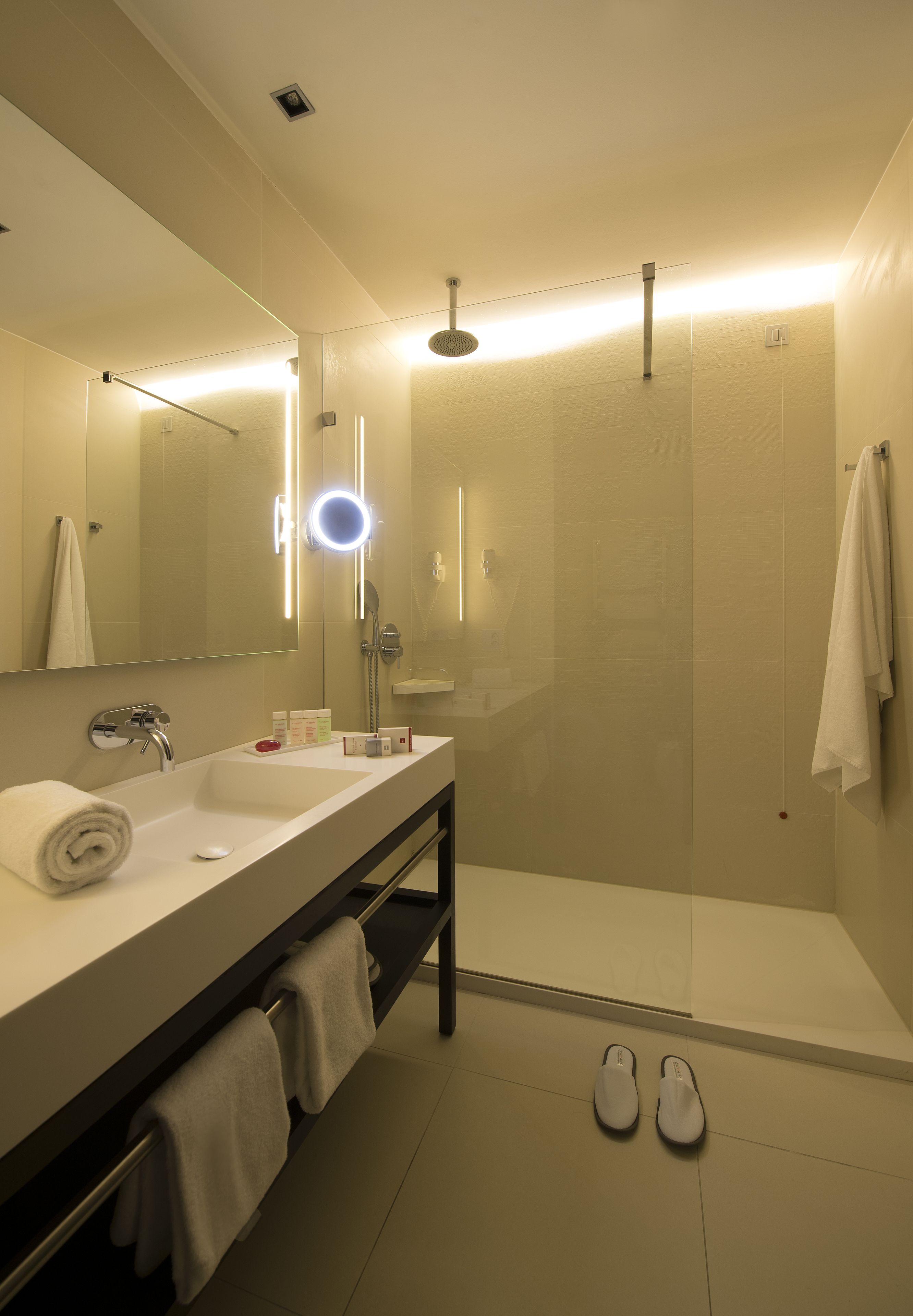 Une salle de bain moderne avec un grand miroir, un lavabo, une douche et une paire de chaussons posés sur le sol.