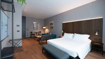 A modern hotel room with a large bed, a seating area with a sofa and chairs, a small table, and a desk.