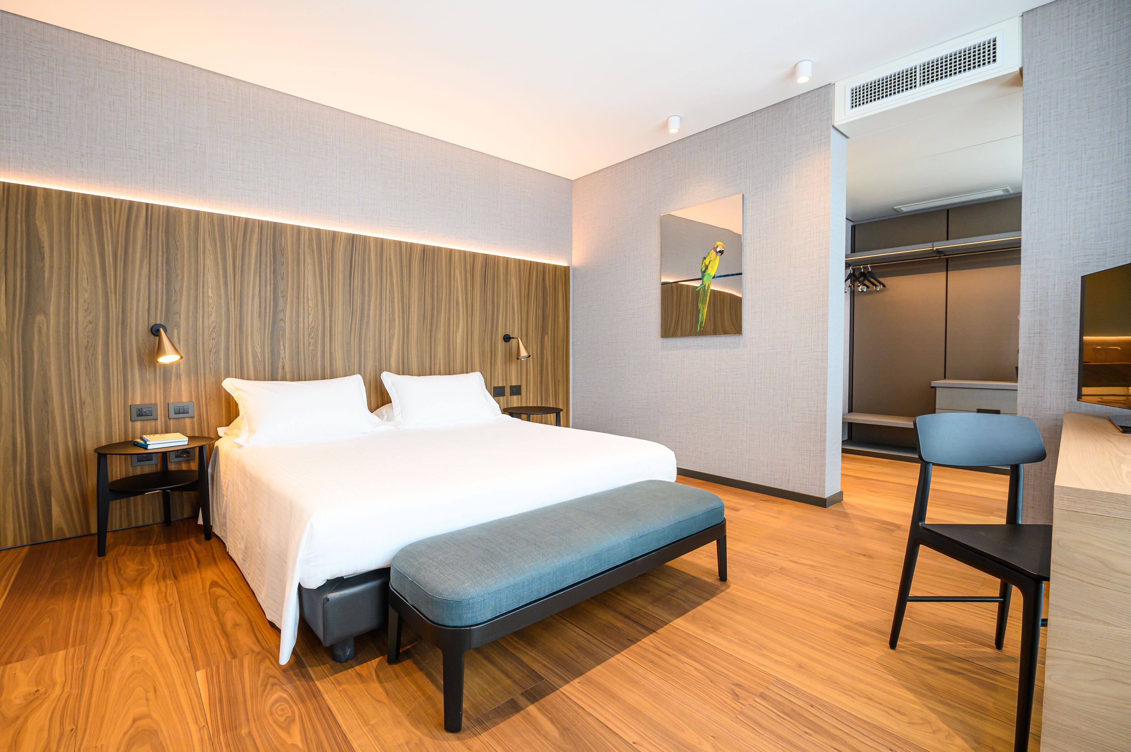 Ein modernes Hotelzimmer mit einem großen Bett, einer Bank, einem Sessel, einem Nachttisch, einem Spiegel und einem Kleiderschrank.