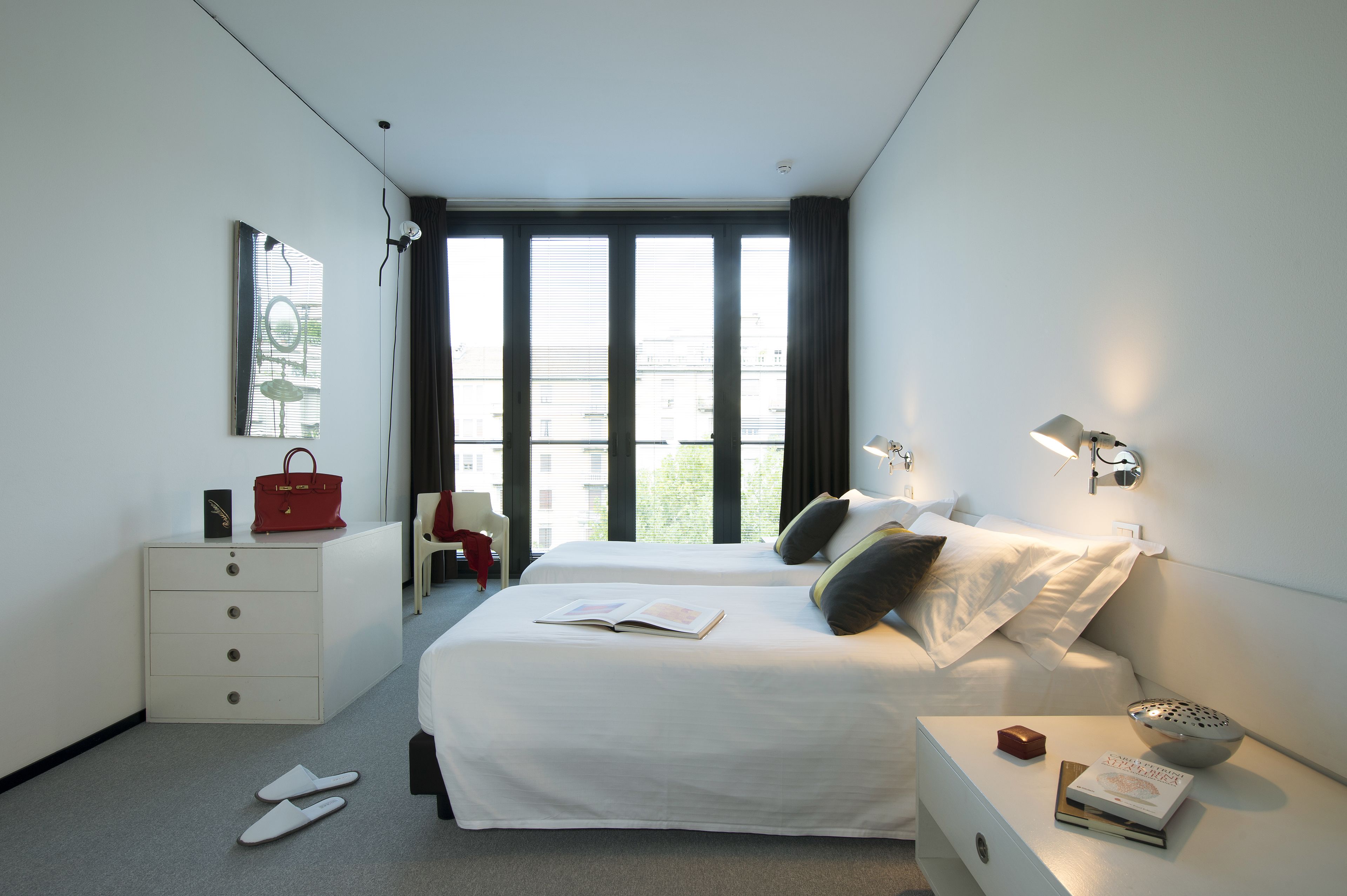 A hotel room with two beds, a white dresser, a red handbag, a chair, a window with blinds, and a nightstand with a lamp and a book.