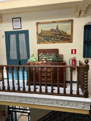 A room with a wooden desk, a framed painting, a blue door, and a fire extinguisher. - Pensión Catedral (Seville)