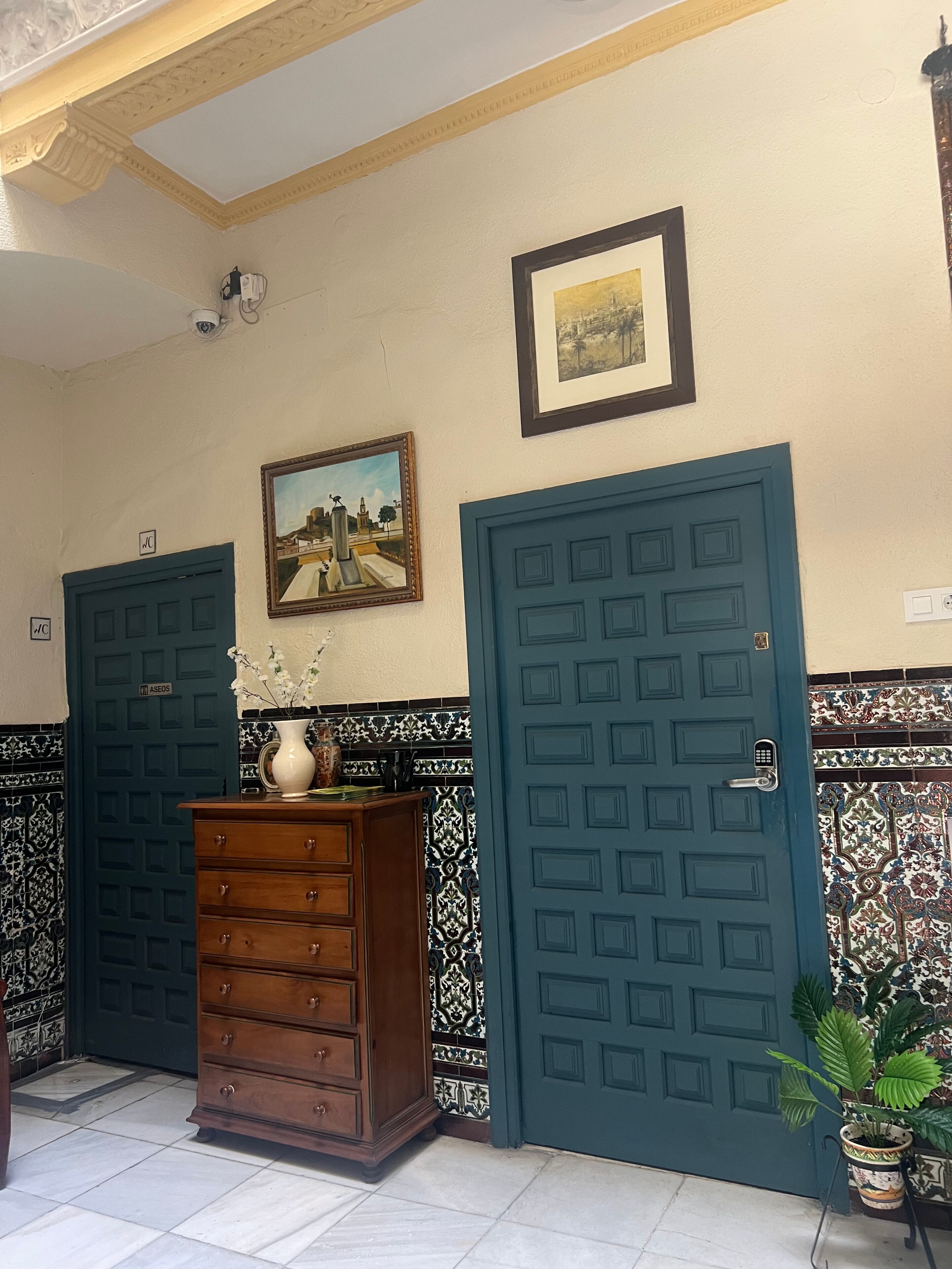 A room with two blue doors, a wooden dresser, a vase with flowers, a framed painting, and patterned tiles on the walls.