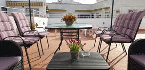 Eine Dachterrasse mit einem Glastisch, umgeben von gestreiften Stühlen, einem Blumentopf und einer klaren Aschenbecher.