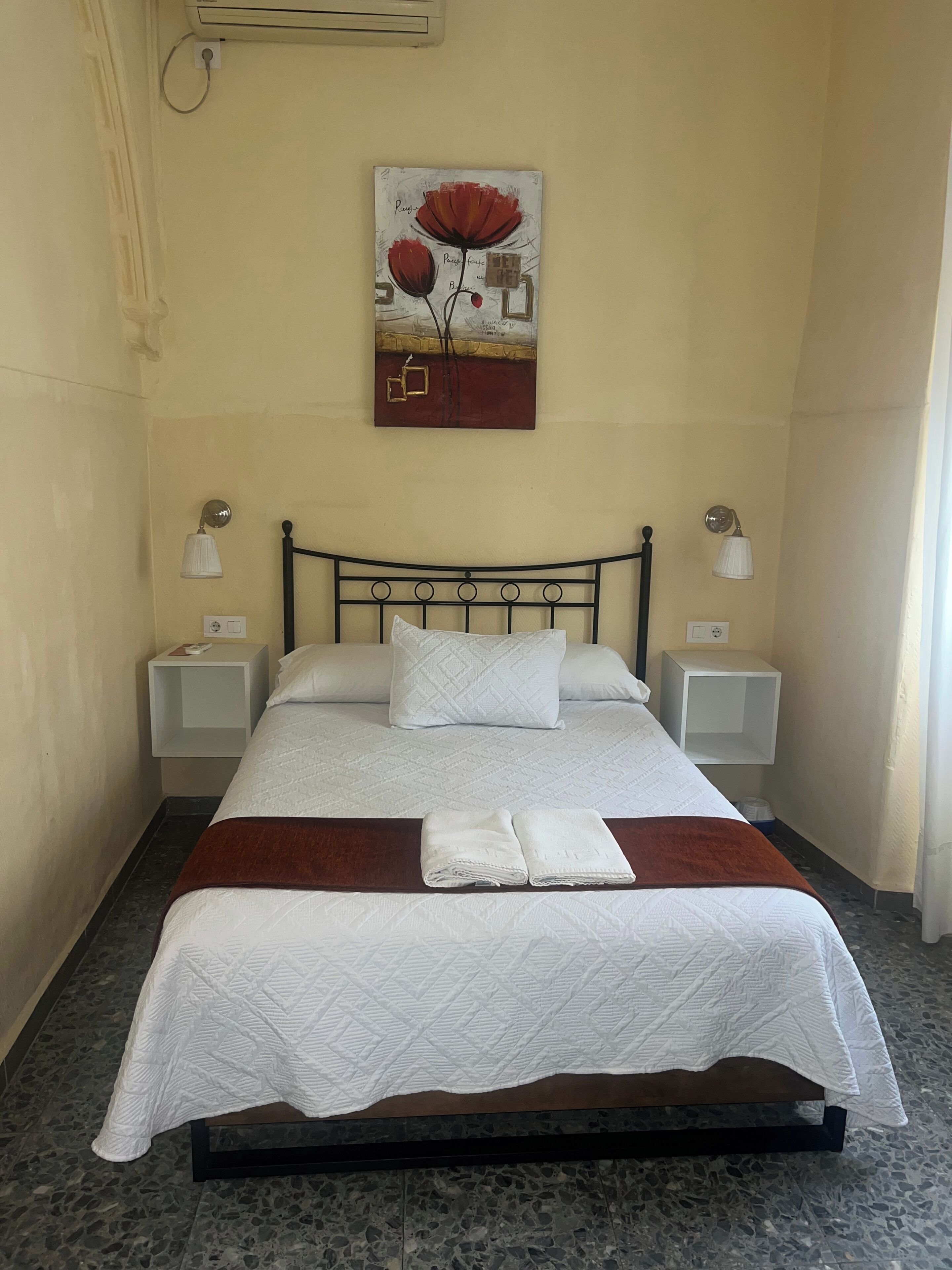 A neatly made bed with white linens, a brown throw blanket, and a painting above it. There are two bedside tables with lamps, and the room has a tiled floor.