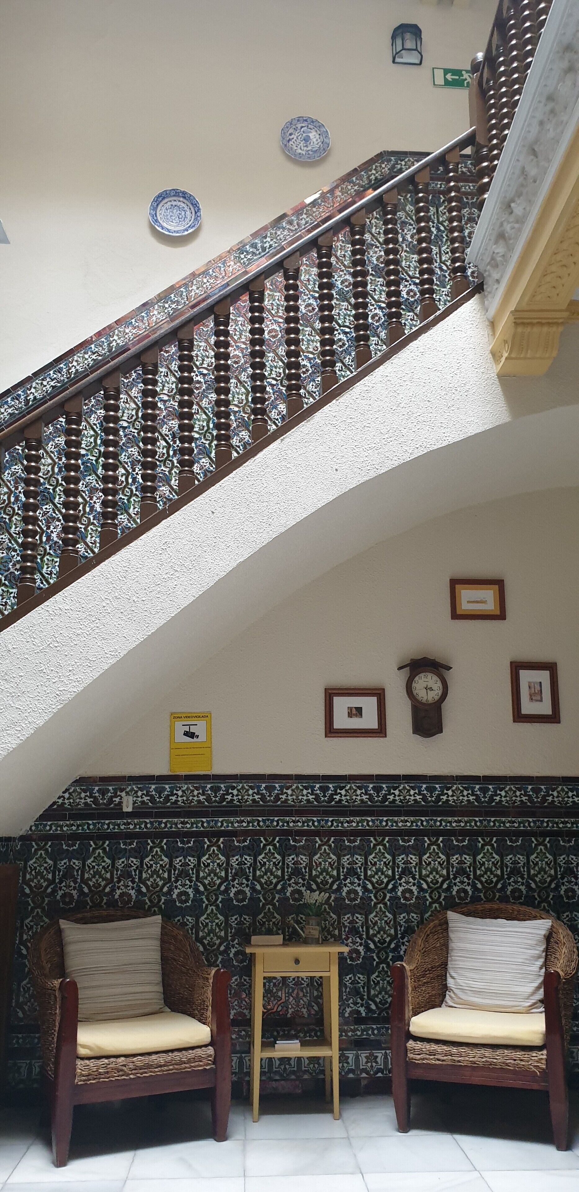 Un escalier orné de balustres travaillés, une horloge, des tableaux encadrés et deux fauteuils en osier avec des coussins.