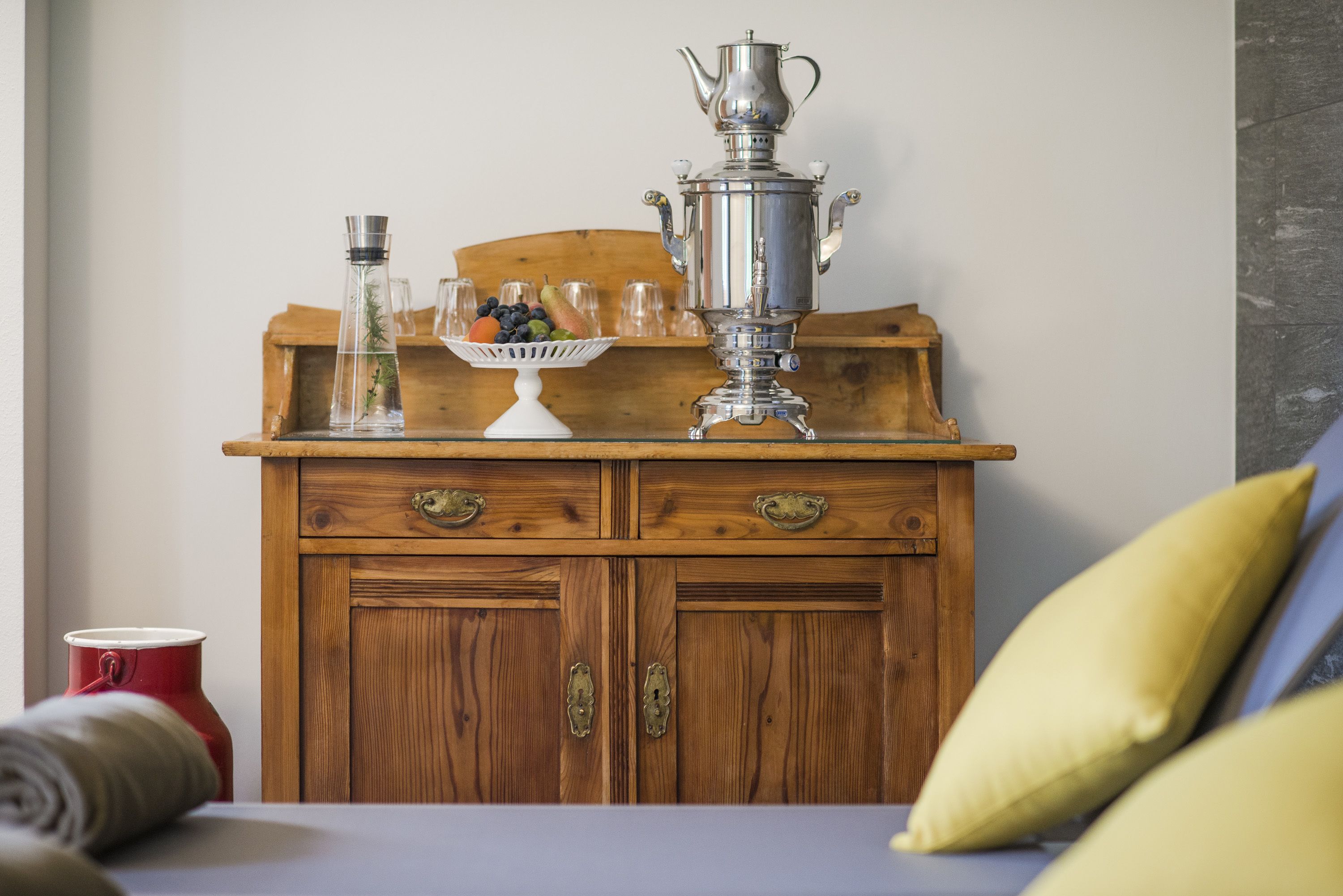 A wooden dresser with two drawers, a metal teapot, and a glass bowl with fruits.