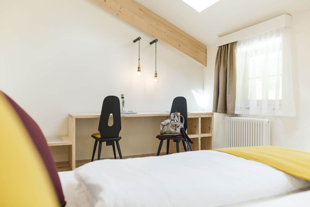 Un dormitorio con cama, escritorio, dos sillas, radiador y ventana con cortinas.