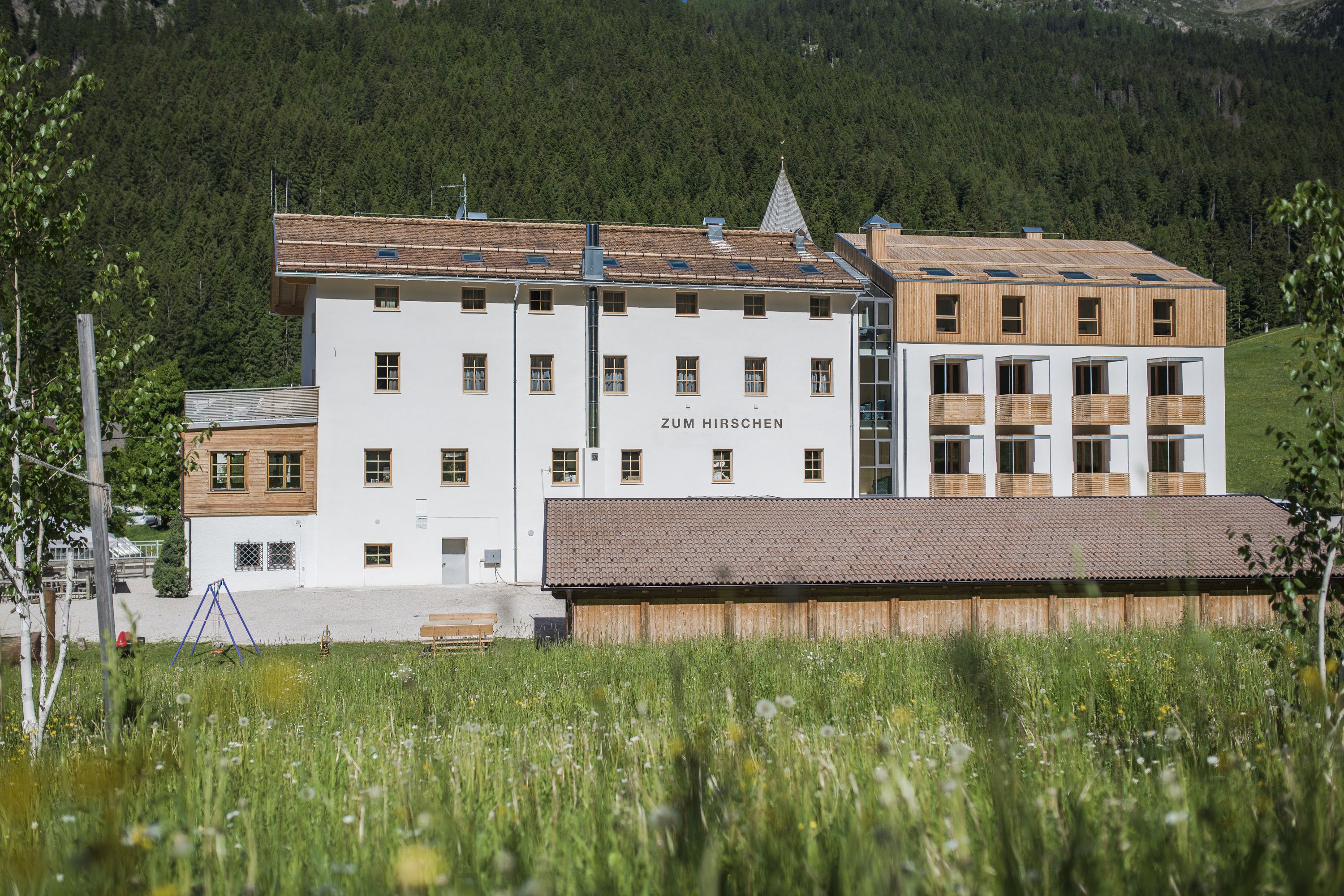 Un edificio a più piani con la scritta "Zum Hirschchen" sulla facciata, immerso in un prato verdeggiante e circondato da dolci colline ricoperte di boschi.