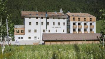 Un edificio a più piani con la scritta "Zum Hirschchen" sulla facciata, immerso in un prato verdeggiante e circondato da dolci colline ricoperte di boschi.