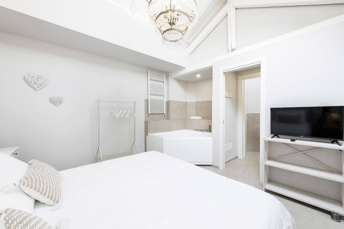 A modern hotel room with a large bed, a TV on a stand, a bathtub, and a chandelier.