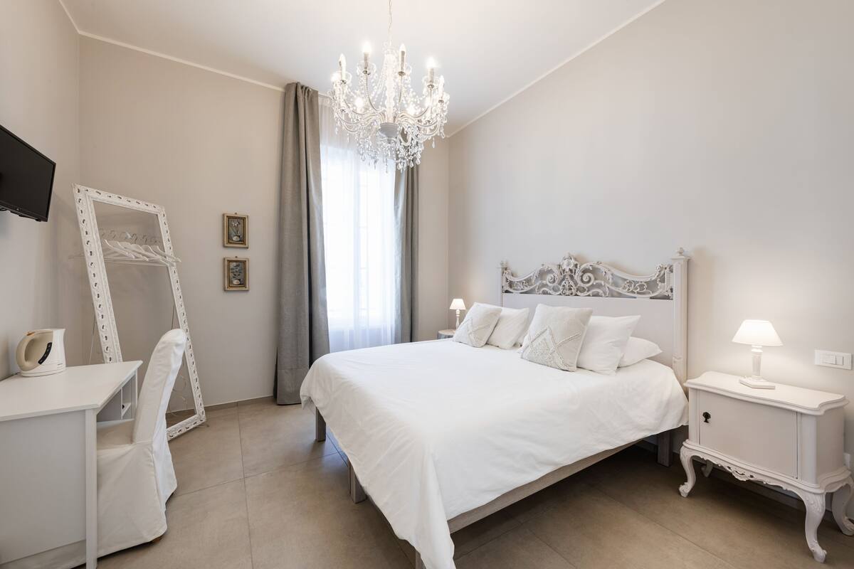 A bedroom with a large bed, a desk, a chair, a dresser, a TV, and a chandelier.
